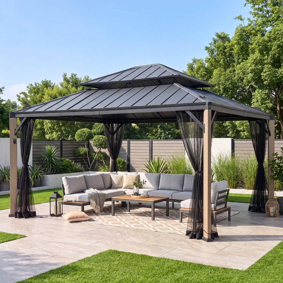 A large outdoor double-roof gazebo with black mesh netting is set on a paved patio in a landscaped backyard. Underneath the gazebo is a gray sectional sofa, coffee table, and rug, surrounded by green grass, shrubs, and trees under a clear blue sky.