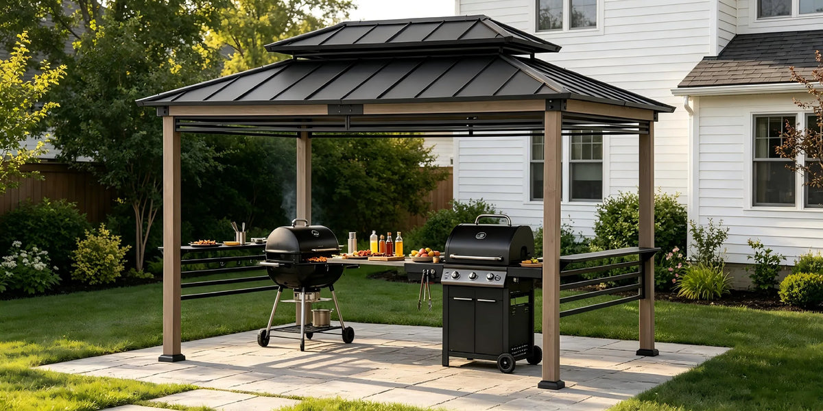 A double-tiered metal gazebiover with a black roof and brown posts, set on a stone patio in a backyard next to a white house. Underneath the cover, a black charcoal grill and a gas grill are positioned on a side table with drinks and food.